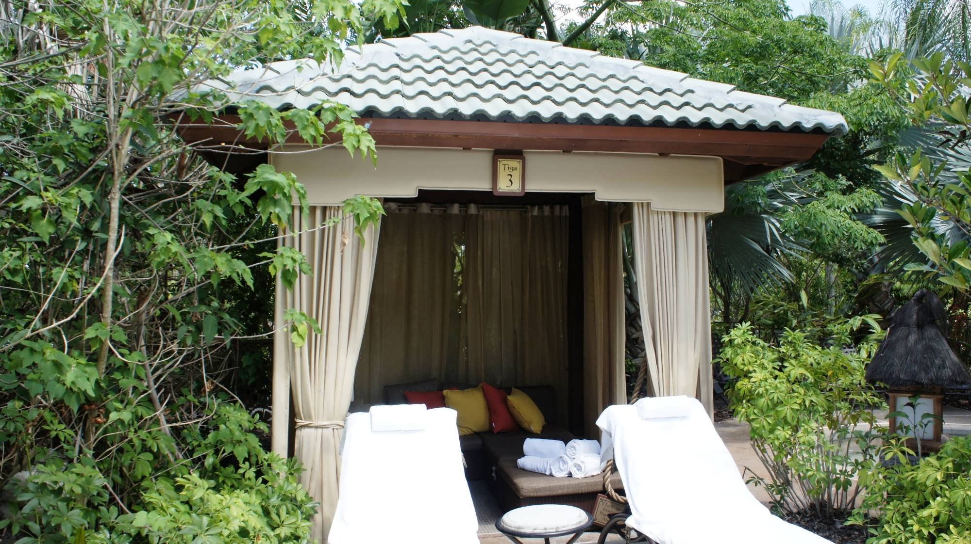 A cabana with curtains, two lounge chairs, and a table tucked into the lush foliage of Royal Pacific Resort's pool area
