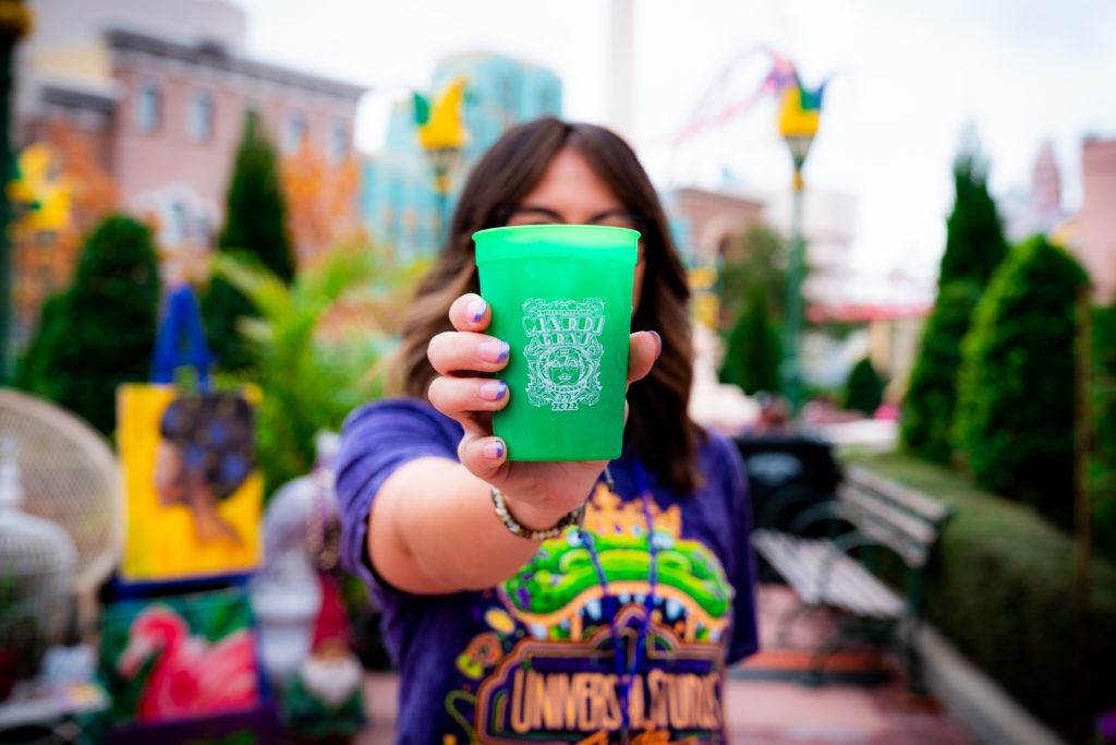 A green plastic cup with a white decal image that includes the words "Universal Orlando's Mardi Gras 2022", held out by a hand with purple tipped nails.