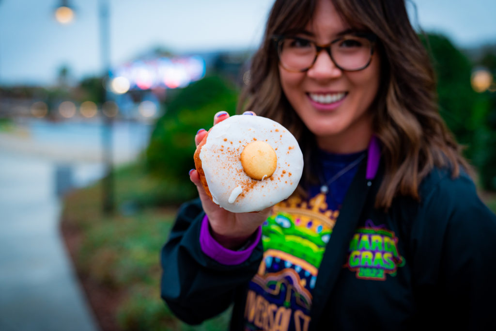 A golden brown circular doughnut shell topped with white icing, cinnamon, and a Nilla wafer, held up by a hand with purple tipped nails. 