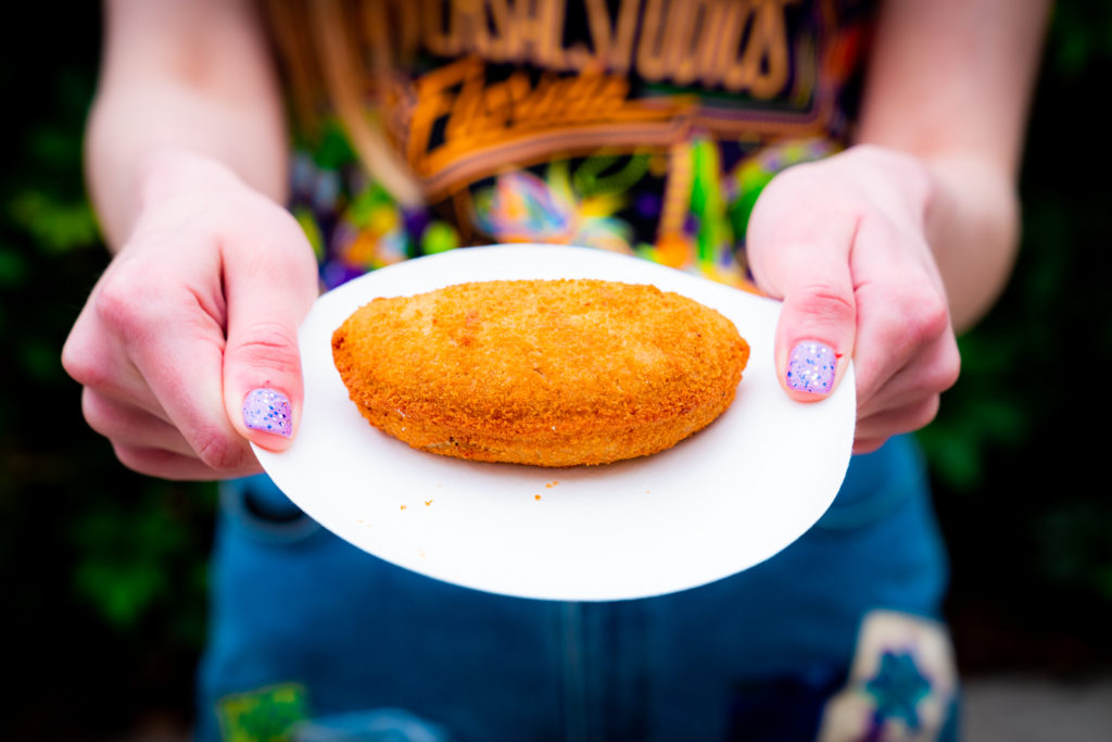 Golden brown, breaded, flat, circular rissole on a white plate, held out by two hands with light purple glitter nail polish. 