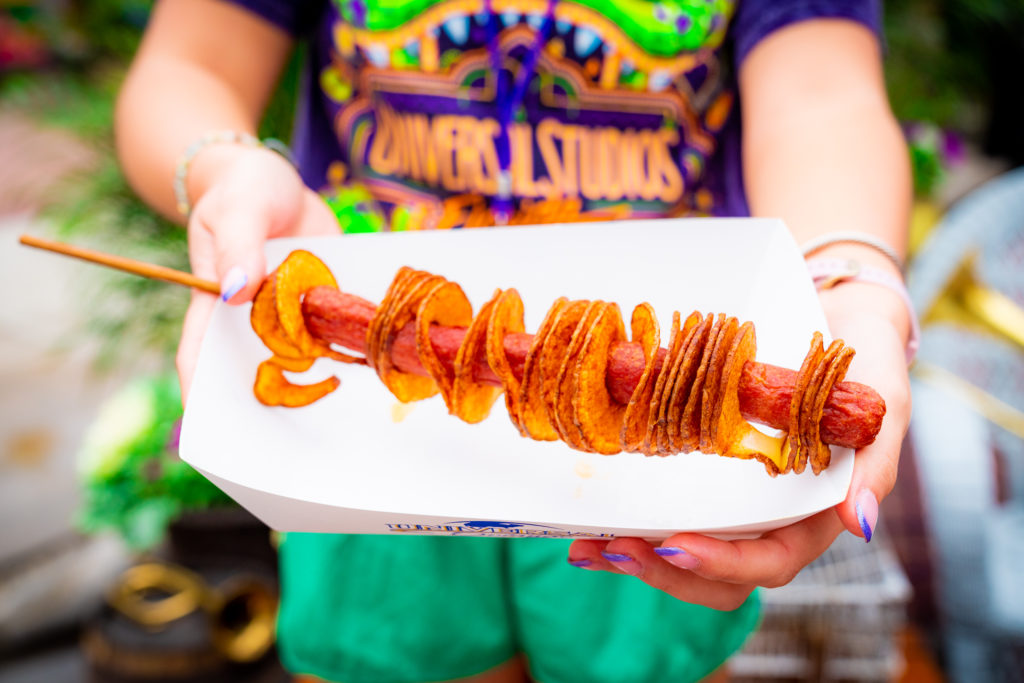 A foot-long hot dog on a skewer wrapped in fried ribbon potato slices in a white paper serving boat, held out by two hands featuring purple tipped nails. 