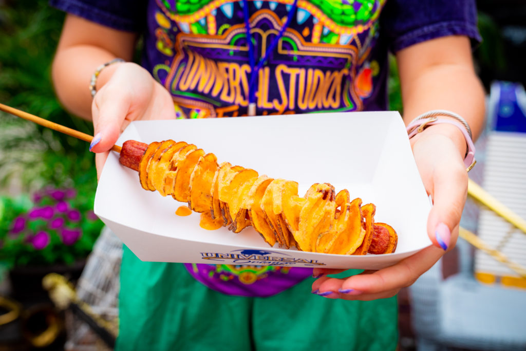 A foot-long hot dog on a skewer wrapped in fried ribbon potato slices and topped with yellow queso in a white paper serving boat, held out by two hands featuring purple tipped nails. 