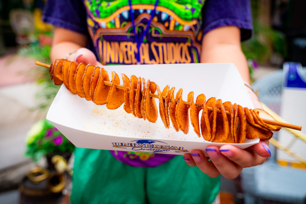 Fried ribbon potato slices on a wooden skewer in a paper serving boat with Universal Orlando's logo on the front, held out by two hands featuring purple tipped nails. 