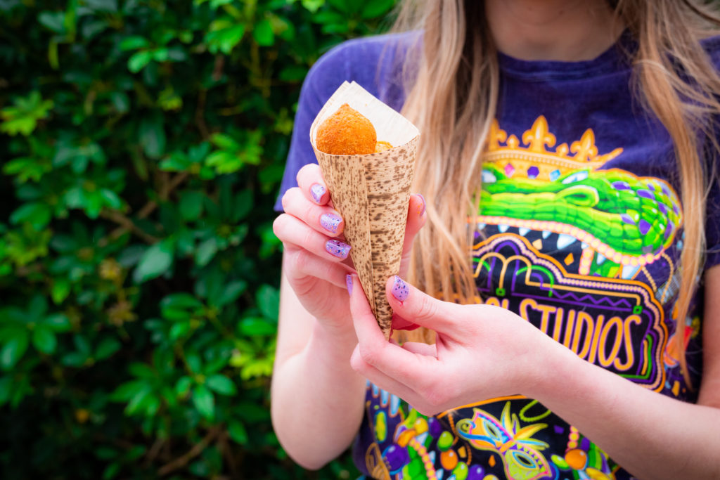 Golden brown fritter balls in a tan and brown paper cone, held by two hands with light purple glitter nail polish. 