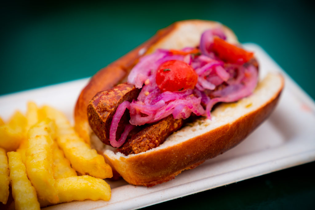 A split-top roll with halved grilled chorizo, garlic mayo, chimichurri, and tomato and red onion curtido next to golden crinkle cut fries on a white rectangular plate. 