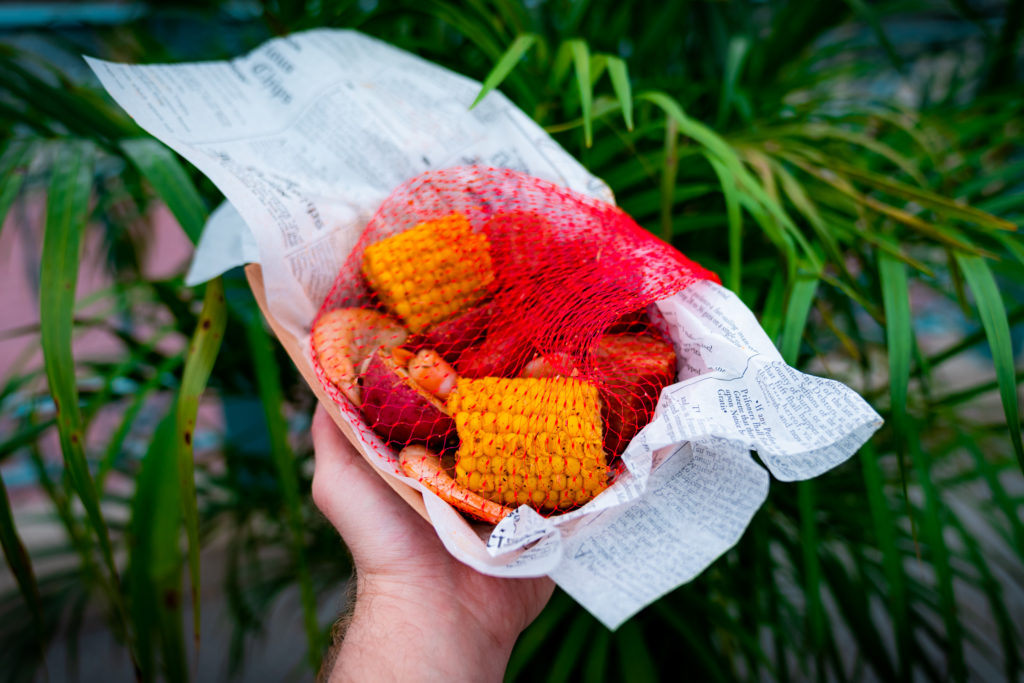 Crawfish, shrimp, andouille sausage, potatoes, and corn on the cob in a red net on top of newspaper in a paper serving boat, held out by a hand. 