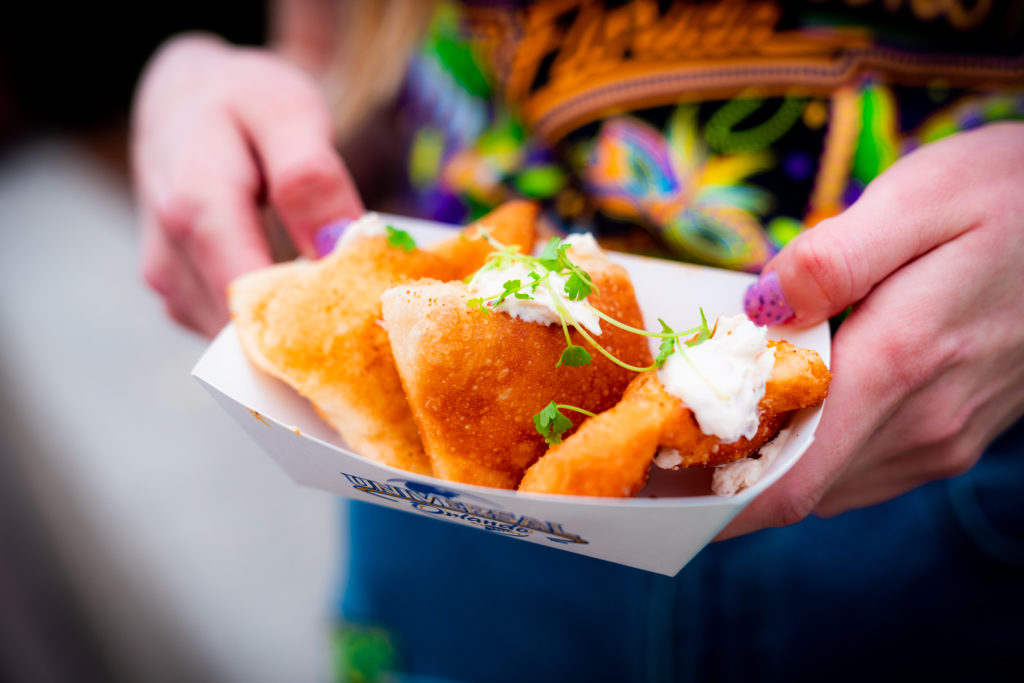 Three beignets dusted with cajun brown sugar, stuffed with crab dip and scallions in a white paper serving bowl with Universal Orlando's logo, held by two hands with light purple glitter nail polish. 