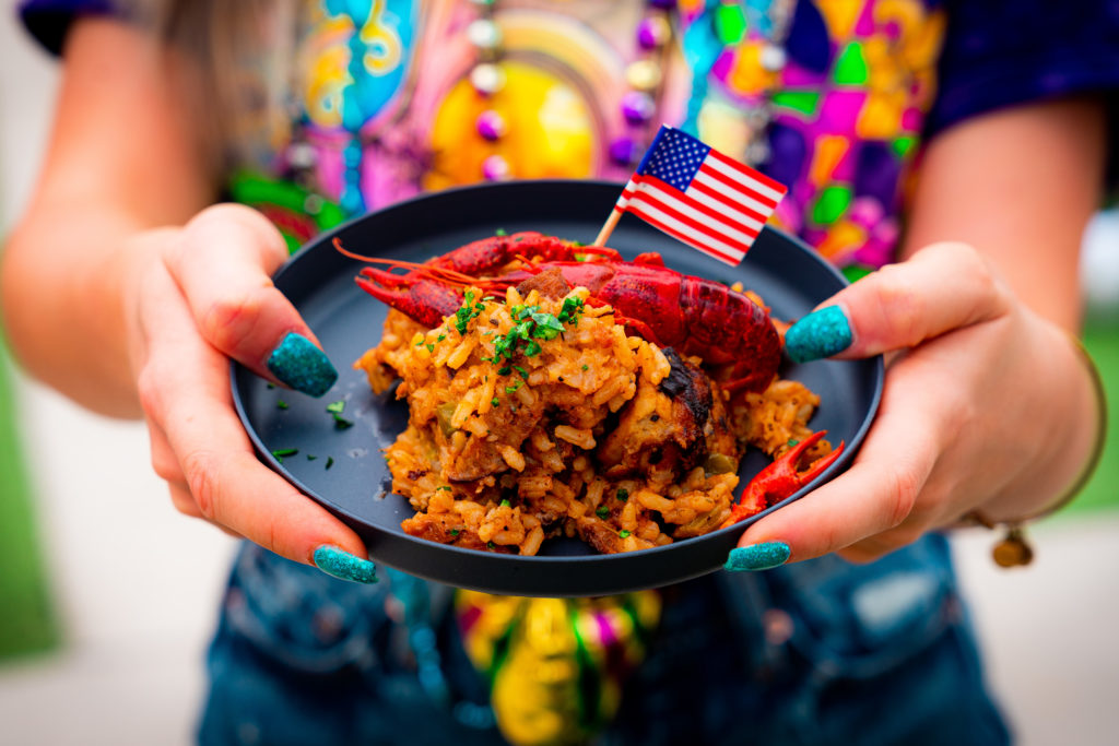 Cajun spiced rice with sausage, chicken, shrimp, tomatoes, peppers, and garnished with a crawfish with a small American flag decoration, on a black plate, held out by two hands with blue sparkle nails. 