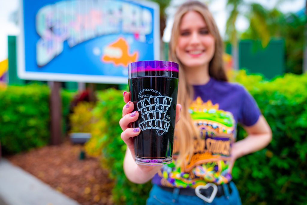 A dark purple plastic cup with a white decal image of beads and the words "Party out of this world!", held forward by a hand with light purple glitter nails. 