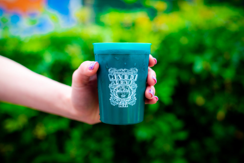 A dark green plastic cup with a white decal image featuring the words "Universal Orlando's Mardi Gras 2022", held by a hand with light purple glitter nails. 