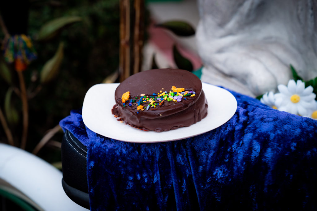 A MoonPie like dessert sandwich made with graham cracker cookies filled with fresh marshmallow covered in chocolate and topped with blue, green, yellow, purple, and gold Mardi Gras sprinkles on a white plate. 
