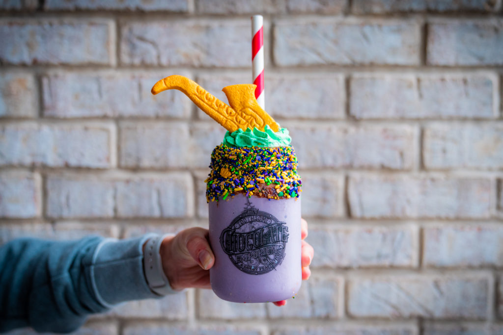 A purple milkshake in a clear plastic jar with Nutella spread generously around the rim with yellow, green, purple, and blue Mardi Gras sprinkles, green whipped topping with a cookie saxophone, and a red and white striped straw. The cup, held out by a hand, has a black decal image reading "Toothsome Chocolate Emporium & Savory Feast Kitchen". 