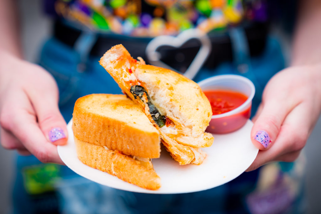 A Texas Garlic Toast sandwich cut in half with one half presented on top of the other containing meatballs and cheese, with a side of red sauce in a small plastic container on a white plate, held out by two hands with light purple glitter nails. 