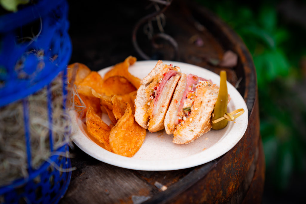 Two sandwich triangles containing mortadella, salami, ham, mozzarella, provolone, and olive relish, on sesame seed buns, skewered with okra and kettle chips on the side on a white plate. 