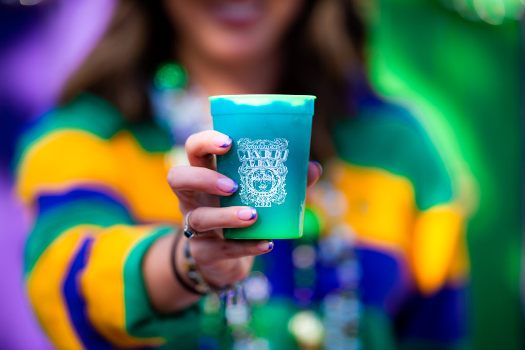 Cup with different shads of green and a white decal image that includes the words "Universal Orlando's Mardi Gras 2022", held out by a hand with purple tipped nails and a silver ring with blue stone. 
