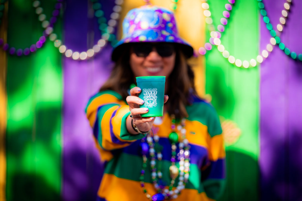 A cup with different shades of green, with a white decal image featuring the words "Universal Orlando's Mardi Gras 2022", held forward by a hand with purple tipped nails and a silver ring with blue stone. 