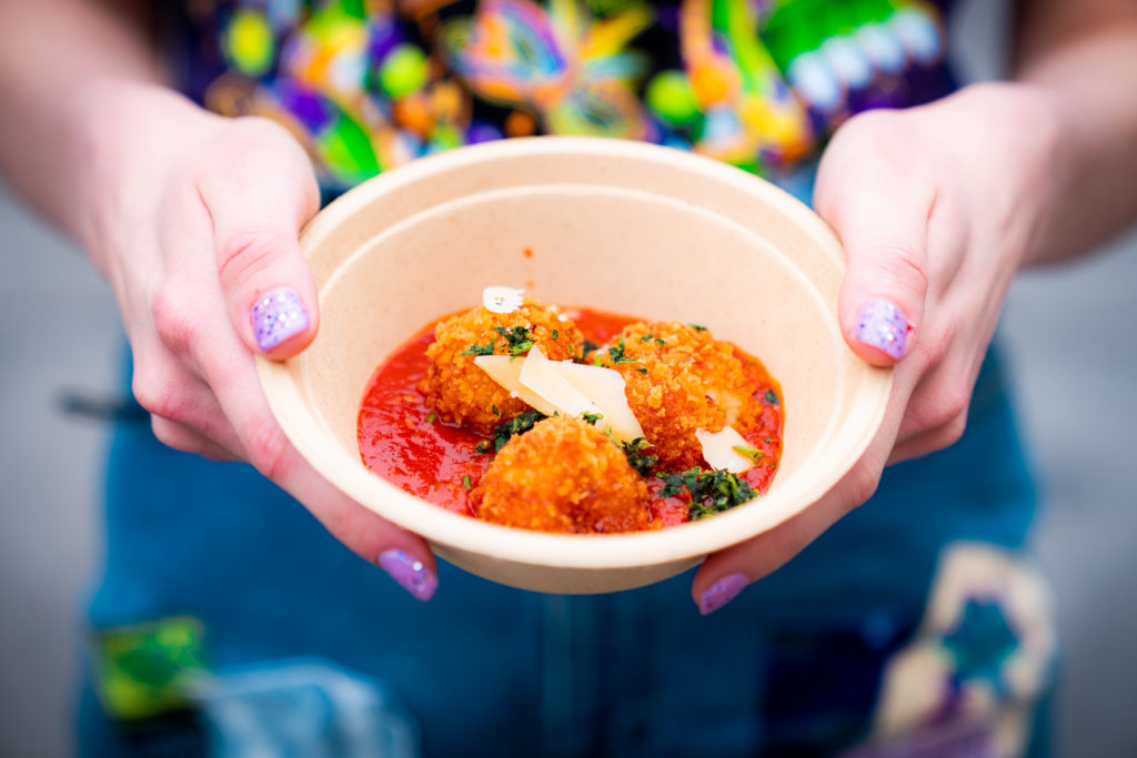 Three fried risotto balls in a red sauce  topped with garnish and cheese in a white bowl, held out by two hands with light purple glitter nails. 