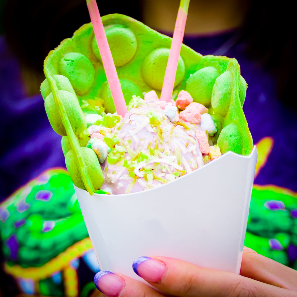 A green bubble waffle with purple ice cream, white coconut shavings, green popping candy, two pink Pocky sticks, and pink, blue, and yellow marshmallows in a white paper cone, held by a hand with purple tipped nails. 