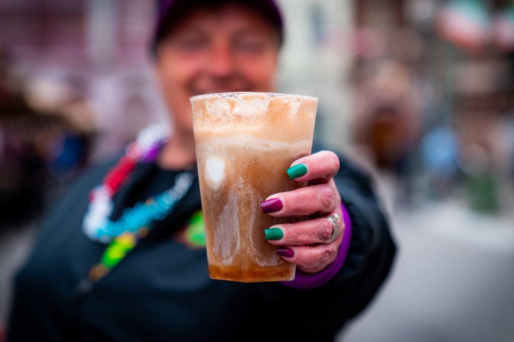 Brown and white swirled drink in a clear plastic cup, held out by a hand with a wedding ring and green and purple alternating nails. 