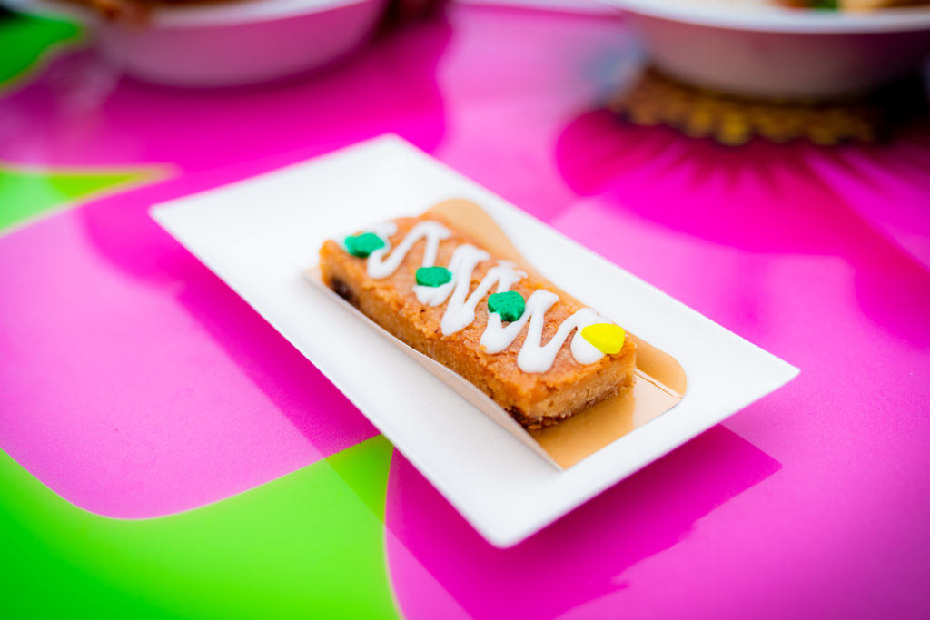 A golden brown pastry bar with white drizzle and green and yellow chocolate pieces in a paper sleeve on a white rectangular plate. 