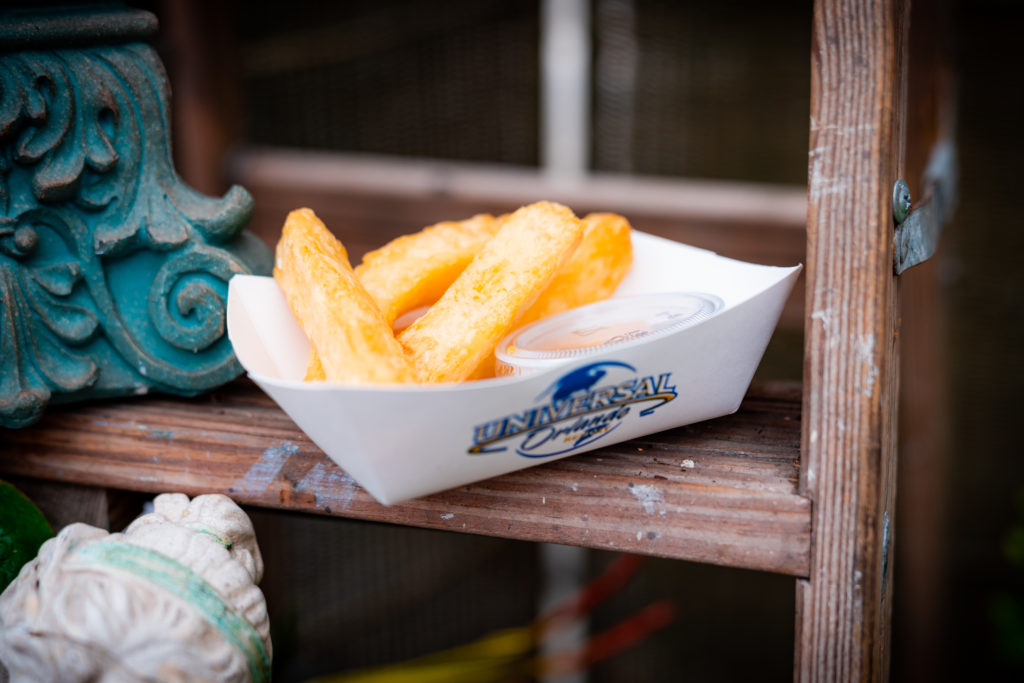Thick cut yucca fries and a plastic sauce container with a lid in a white paper serving boat with Universal Orlando's logo. 